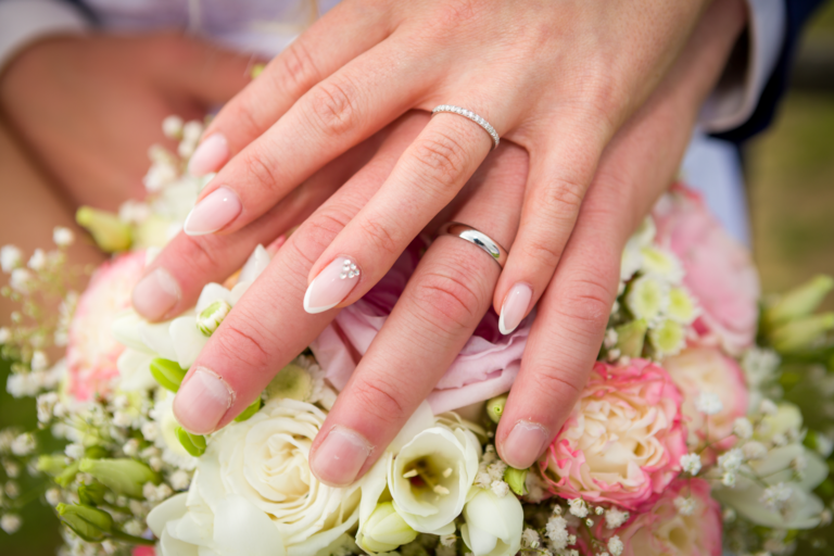 Detail rukou novomanželů s&nbsp;prstýnky položených na&nbsp;svatební kytici